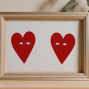 以下が含まれることがあります： 白い背景に、刺繍された目を持つ2つの赤いハートの形が特徴の額装されたアートワーク。 アートワークは金色のフレームに入っており、木製の表面に表示されています。