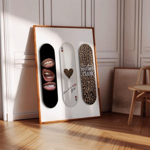May include: Three skateboard decks displayed in a wooden frame. The first deck is black with gold teeth and lips graphics. The second is white with a leopard print heart and text. The third deck has a leopard print pattern and the text "Bitches Club".