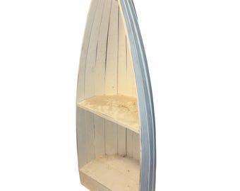May include: A tall, vertical wooden shelf shaped like a boat, featuring a light blue exterior and a white interior. The structure includes two horizontal shelves for storage, with a pointed top and a flat base, constructed from vertical wooden planks.