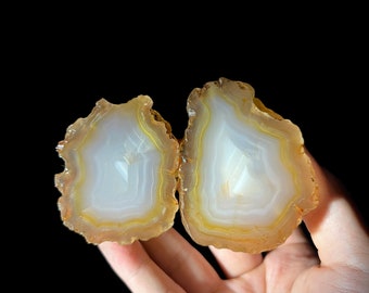 May include: Two halves of a geode with white and yellow agate banding. The geode is held in a hand.