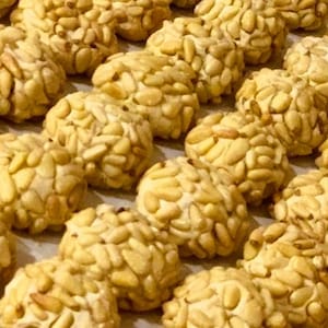 May include: Close-up of a tray of small, round, golden-brown cookies covered in pine nuts. The cookies are arranged in rows and are slightly overlapping.