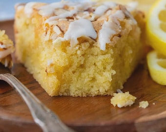 May include: A slice of lemon cake with white icing and almond slivers on a wooden plate. The cake is yellow and has a textured appearance. Lemon slices are visible in the background, along with a silver fork.