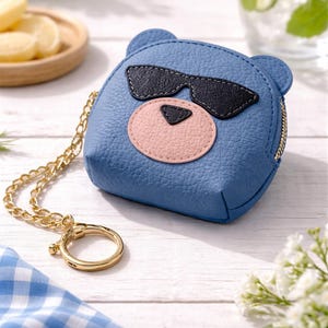 May include: A blue coin purse shaped like a bear's head, with black sunglasses and a pink nose. It has a gold chain and ring. The purse is on a white wooden surface, with a blue and white checkered cloth in the foreground.