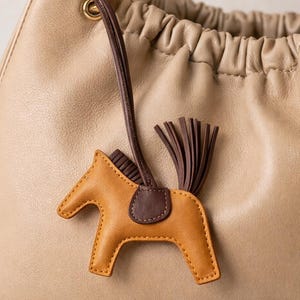 May include: A tan leather handbag with a brown leather strap and gold-tone hardware. A small, tan and brown leather horse charm with a dark brown saddle and tail hangs from the bag's handle. The bag has a gathered top.