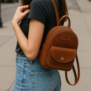 May include: A brown leather backpack with a front pocket and a top handle. The backpack has a rounded shape and visible stitching. The straps are adjustable. The person is wearing a dark grey t-shirt and blue jeans.