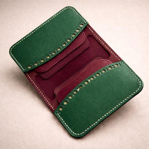 Puede incluir: Cartera de cuero abierta en verde y burdeos. La cartera tiene un diseño de dos pliegues con múltiples ranuras para tarjetas. Los bordes están cosidos con hilo blanco y las solapas verdes tienen perforaciones decorativas. El interior es burdeos.