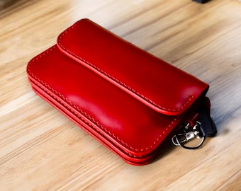 May include: A red leather key holder with a flap closure. The key holder is rectangular with rounded edges and a stitched border. A silver-colored key ring and a black strap are visible.