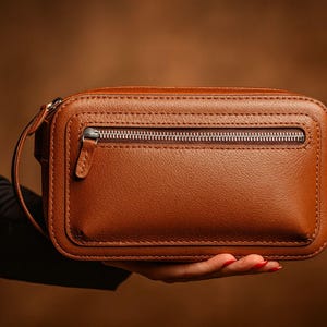 Op de afbeelding: Een rechthoekige bruine leren clutch met een zilveren ritssluiting. De clutch heeft een klein handvat aan de zijkant en wordt in de hand van een persoon gehouden. Het leer lijkt aan de randen gestikt te zijn.