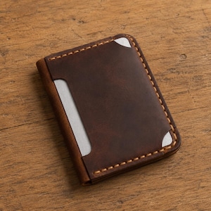 Puede incluir: Un tarjetero de cuero marrón oscuro con costuras visibles de color claro a lo largo de los bordes. La cartera tiene una forma rectangular con esquinas redondeadas e incluye una pieza de metal plateado visible a través de las aberturas frontales.
