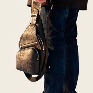 Peut inclure: Un sac &agrave; bandouli&egrave;re en cuir marron avec une boucle argent&eacute;e et des accents de fermeture &eacute;clair. Le sac est port&eacute; sur l'&eacute;paule d'une personne portant un jean bleu.