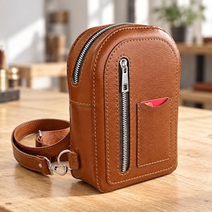 May include: A tan leather crossbody bag with a rounded top, a vertical silver zipper closure, and an exterior slip pocket containing a red card. The bag features a matching adjustable leather strap with silver metal hardware clips.
