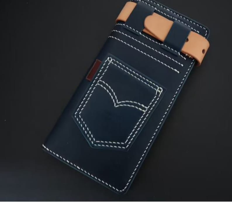 May include: A navy blue leather wallet with a white stitched pocket detail, resembling a classic jean pocket. The wallet has a tan leather strap with a buckle closure.