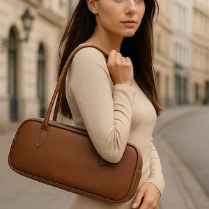 Puede incluir: Un bolso de mano de cuero marrón con forma rectangular y asas largas. El bolso se lleva al hombro. La persona lleva un vestido beige de manga larga.