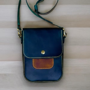 Może przedstawiać: Torebka skórzana w kolorze ciemnoniebieskim z klapką ze skóry brązowej i żółtymi szwami. Torebka ma złoty metalowy zamek i pasek.