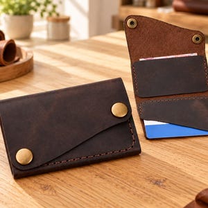 May include: A dark brown leather wallet featuring two brass snap buttons on the front flap. The interior displays two card slots, one of which holds a blue and white card, and two additional brass snap buttons at the top corners.