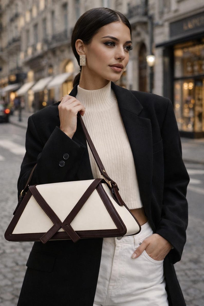 Peut inclure: Sac &agrave; main g&eacute;om&eacute;trique cr&egrave;me et marron fonc&eacute;. La personne porte un blazer noir, un col roul&eacute; cr&egrave;me et un jean blanc. En arri&egrave;re-plan, une rue de la ville avec des b&acirc;timents.