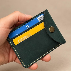 May include: A dark green leather card holder featuring a rounded flap secured with a brass-colored snap button. The wallet has visible light-colored stitching along the edges and contains two visible cards, one blue and one yellow with the text aixess.