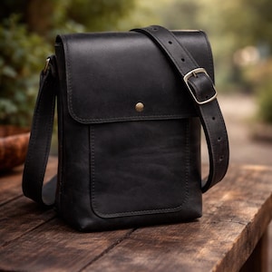 Pode incluir: Bolsa mensageiro de couro preto com fecho de aba e alça de ombro ajustável. A bolsa possui um bolso frontal e um fecho de botão cor de latão. A bolsa está sobre uma superfície de madeira.