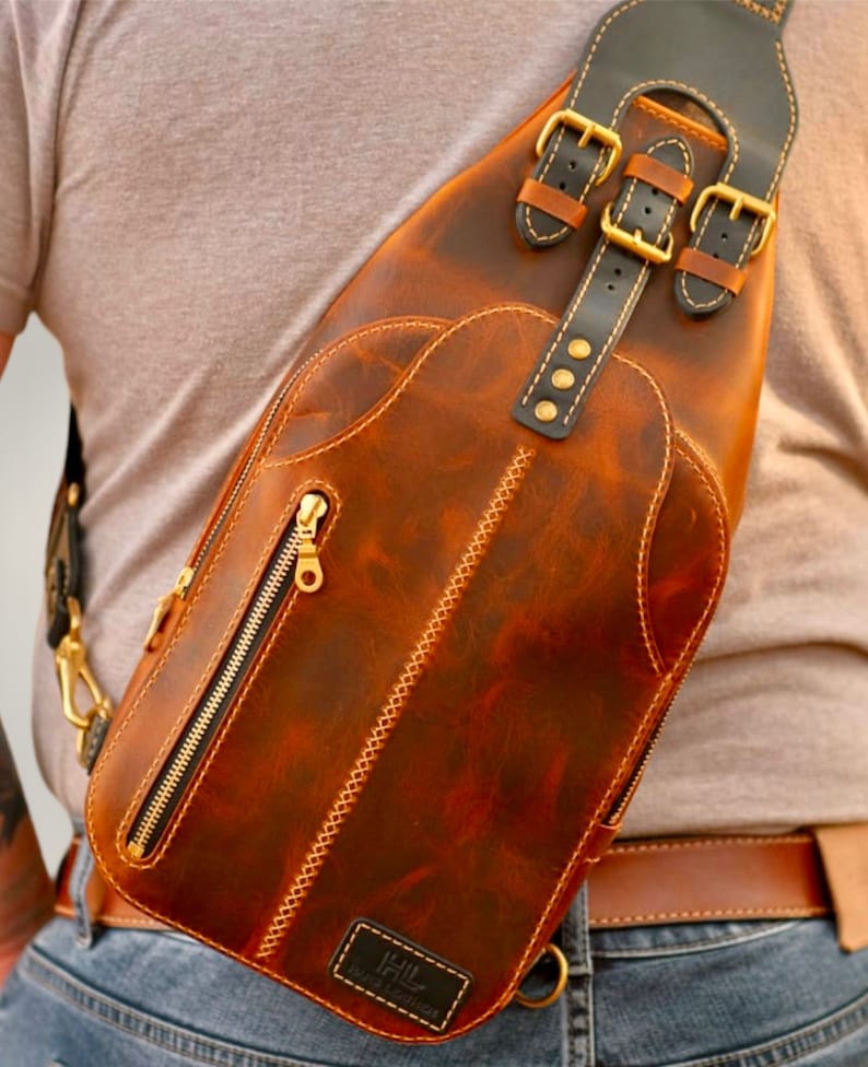 May include: A brown leather crossbody bag with a black strap and gold buckles. The bag has a zipped pocket on the front and a small leather patch with the text "Hill Handcrafted" on the bottom.