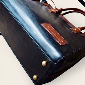 Pode incluir: Bolsa de m&atilde;o de couro preto com detalhes em couro marrom, com formato retangular e tachas de metal dourado na base. A bolsa tem uma al&ccedil;a superior e uma al&ccedil;a de ombro. Uma etiqueta de couro marrom &eacute; costurada na frente.
