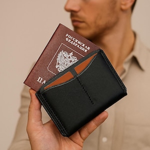 Könnte beinhalten: Schwarzes Lederportemonnaie mit braunem Innenraum, das einen weinroten Reisepass enthält. Der Reisepass hat kyrillischen Text und ein Wappen. Das Portemonnaie hat sichtbare Nähte und ein minimalistisches Design.