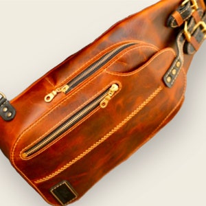 May include: A brown leather crossbody bag with two zipped compartments and a gold buckle strap. The bag has a distressed look and is made of high-quality leather.