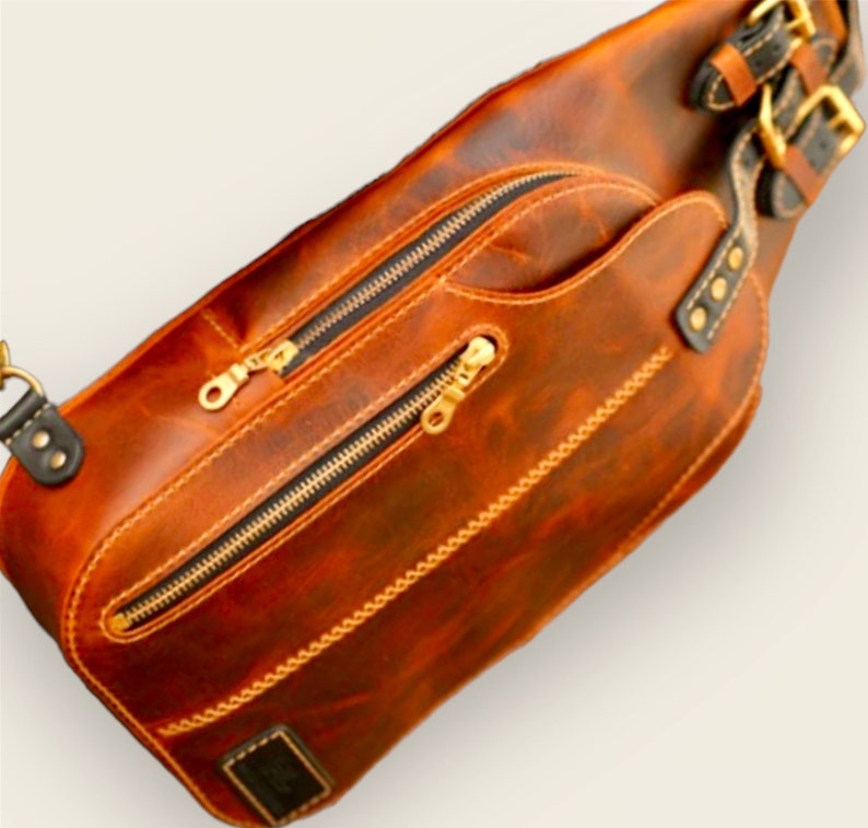 May include: A brown leather crossbody bag with two zipped compartments and a gold buckle strap. The bag has a distressed look and is made of high-quality leather.