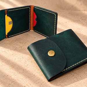 Puede incluir: Una cartera de cuero verde oscuro con una solapa redondeada y un cierre de botón a presión de latón. La cartera presenta costuras amarillas en contraste a lo largo de los bordes. Una cartera se muestra cerrada, mientras que otra está abierta, revelando ranuras para tarjetas con una tarjeta amarilla y una roja.