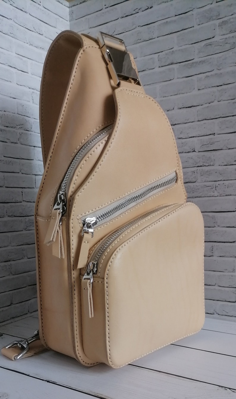 Peut inclure: Un sac &agrave; bandouli&egrave;re en cuir beige avec une boucle argent&eacute;e et plusieurs compartiments zipp&eacute;s.