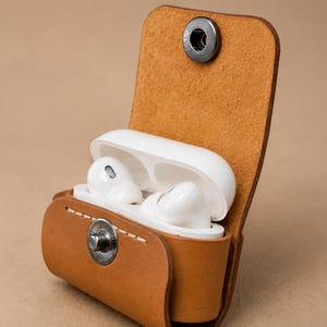 Puede incluir: Un estuche de cuero color canela con cierre de botón a presión metálico que contiene un par de auriculares inalámbricos blancos. El estuche presenta costuras visibles a lo largo de la solapa frontal y mantiene la unidad de carga en posición vertical.