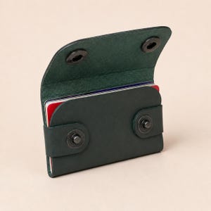 Pode incluir: Um porta-cartões de couro verde-escuro com fecho de aba fixado por dois botões de pressão de metal preto. O painel frontal apresenta uma tira a condizer com dois botões de pressão de metal preto adicionais, contendo vários cartões.