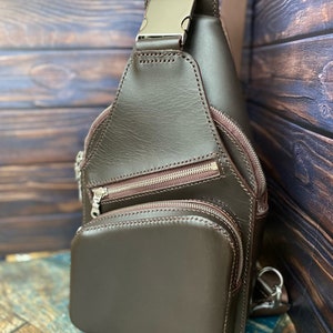 Peut inclure: Un sac &agrave; bandouli&egrave;re en cuir marron avec plusieurs compartiments &agrave; fermeture &eacute;clair et une boucle argent&eacute;e. Le sac est photographi&eacute; sur une surface en bois.