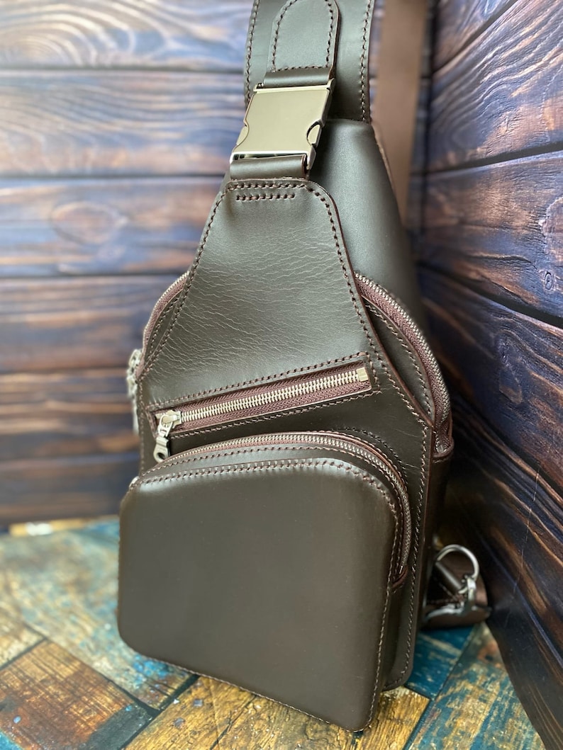 Peut inclure: Un sac &agrave; bandouli&egrave;re en cuir marron avec plusieurs compartiments &agrave; fermeture &eacute;clair et une boucle argent&eacute;e. Le sac est photographi&eacute; sur une surface en bois.