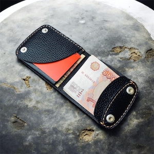 Puede incluir: Cartera de cuero negro con diseño de dos pliegues, con costuras beige en contraste. La cartera está abierta, revelando una tarjeta roja y varios billetes rusos. Dos remaches plateados aseguran la solapa superior.