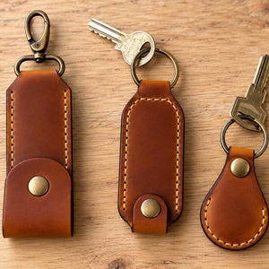 May include: Three brown leather keychains with brass-toned hardware and visible stitching. The left keychain has a lobster clasp, while the center and right keychains feature circular rings, with keys attached to the center and right items.