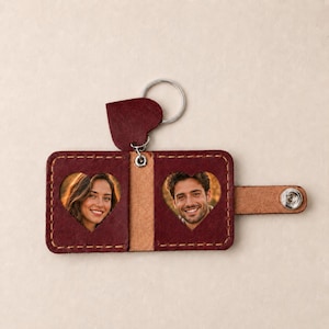 May include: A dark red leather keyring featuring a folding design with a metal snap closure. The interior displays two heart-shaped cutouts, each framing a portrait photograph of a person. A heart-shaped leather charm is attached to the metal ring.