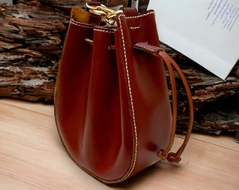 May include: A small, brown leather bucket bag with a drawstring closure. The bag has a rounded base and a gold-colored chain strap. White stitching accents the seams. The bag is approximately 7 inches tall.