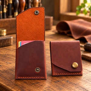 May include: Two dark brown leather card holders with visible stitching. One holder is open, revealing an orange interior and a metal snap button, while the other is closed with a brass-coloured button. Both items feature a slanted front pocket design.