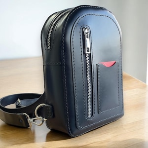 May include: A black leather crossbody bag with a zippered pocket and a red accent. The bag has a strap and a silver buckle.