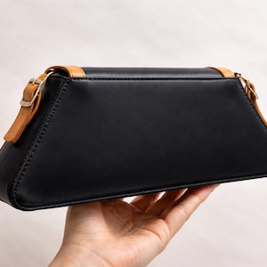 Peut inclure: Un sac &agrave; main en cuir noir de forme trap&eacute;zo&iuml;dale. Le sac est dot&eacute; d'une bandouli&egrave;re en cuir beige et d'une garniture dor&eacute;e. Le sac est tenu dans la main d'une personne sur un fond neutre. Le sac mesure environ 25 cm de large.
