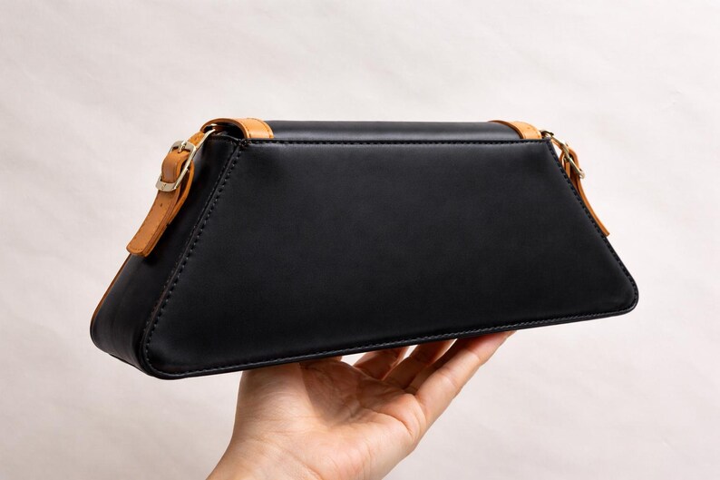 Peut inclure: Un sac &agrave; main en cuir noir de forme trap&eacute;zo&iuml;dale. Le sac est dot&eacute; d'une bandouli&egrave;re en cuir beige et d'une garniture dor&eacute;e. Le sac est tenu dans la main d'une personne sur un fond neutre. Le sac mesure environ 25 cm de large.
