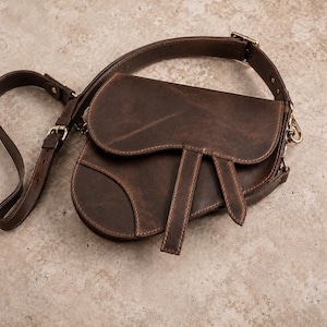 May include: A dark brown leather saddle bag featuring a curved flap, two vertical leather straps hanging from the front, and an adjustable shoulder strap with a metal buckle. The bag has a smooth, matte finish and visible stitching along the edges.