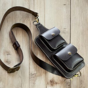 Puede incluir: Un bolso de cuero marrón cruzado con tres bolsillos y una correa larga ajustable. El bolso tiene un diseño cosido y herrajes metálicos.