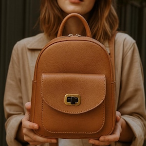 May include: A brown leather backpack with a rounded top and a front flap pocket. The backpack has a top handle and adjustable straps. The flap is secured with a gold-colored clasp. The bag is held by a person.