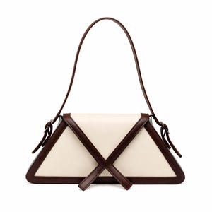 Peut inclure: Un sac &agrave; main cr&egrave;me avec un design g&eacute;om&eacute;trique. Le sac a une forme triangulaire avec des bordures et des accents marron fonc&eacute;. Il poss&egrave;de une longue poign&eacute;e incurv&eacute;e et un motif en forme de croix sur le devant. Le sac est en cuir.
