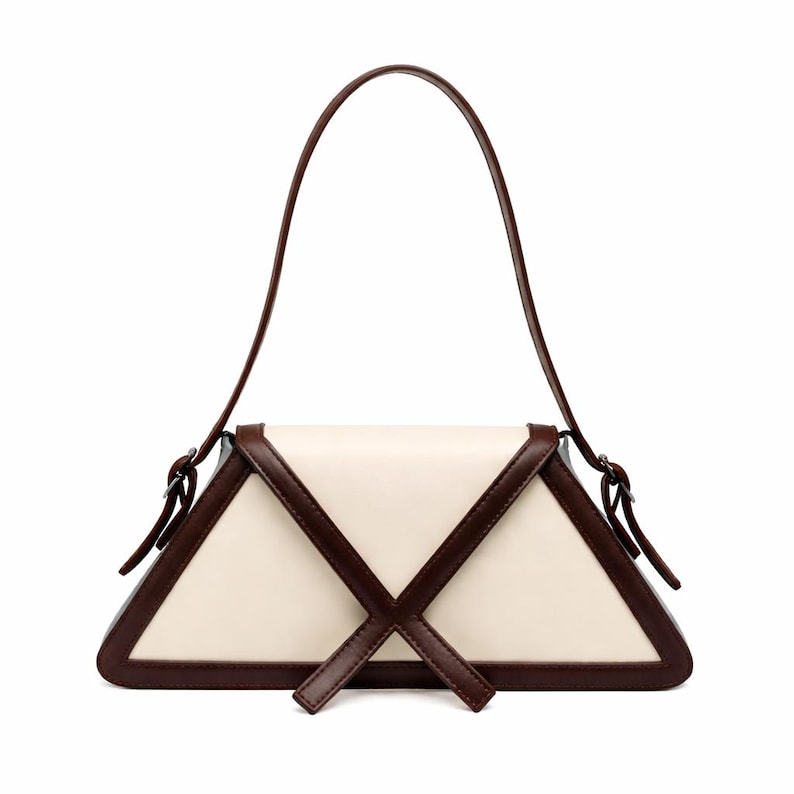 Peut inclure: Un sac &agrave; main cr&egrave;me avec un design g&eacute;om&eacute;trique. Le sac a une forme triangulaire avec des bordures et des accents marron fonc&eacute;. Il poss&egrave;de une longue poign&eacute;e incurv&eacute;e et un motif en forme de croix sur le devant. Le sac est en cuir.
