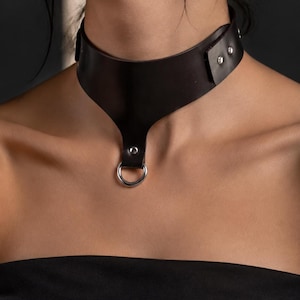 May include: A black leather choker featuring a wide band that tapers to a center point. The design includes a silver-toned metal O-ring attached with a rivet at the bottom and two silver-toned metal snaps on the side for closure and adjustment.