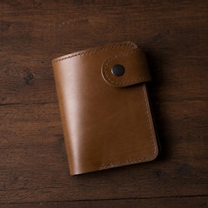 Puede incluir: Cartera de cuero marrón con cierre de botón a presión. La cartera rectangular tiene una superficie lisa y costuras visibles en los bordes. Descansa sobre una superficie de madera oscura.