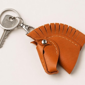 May include: A tan leather keyring shaped like a horse head with a fringed mane, featuring visible light-coloured stitching, a silver-toned metal snap button, and a silver-toned metal split ring with a single silver-toned key attached.