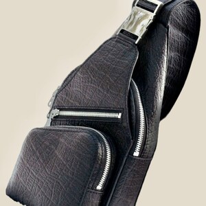 Peut inclure: Un sac &agrave; bandouli&egrave;re en cuir noir avec une boucle argent&eacute;e et des fermetures &agrave; glissi&egrave;re. Le sac a une surface textur&eacute;e et caillouteuse.